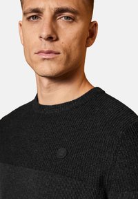 Schwarzer Strickpullover mit einem strukturierten Muster, runder Ausschnitt und kleinem Logo auf der Brust. Glattes Material mit kontrastierenden Texturen.
