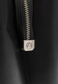 Dettaglio di una borsa in pelle nera con una zip argentata e un tirante inciso con il logo, circondato da un tessuto testurizzato attorno alla cerniera. Superficie liscia.