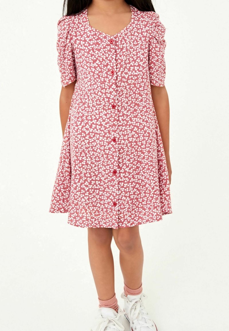 Robe rouge avec un motif de cœurs blancs, manches courtes bouffantes, décolleté en V, devant à boutons. Associée à des baskets blanches et des chaussettes rose pâle.