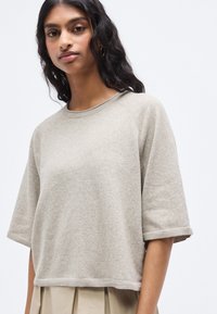 Pull beige à manches courtes en tissu doux et texturé avec un col arrondi et des bords côtelés, offrant une coupe décontractée et carrée.