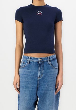 Frau trägt ein marineblaues, kurzes Tommy Jeans T-Shirt mit Logo und hoch taillierte, verblasste blaue Jeans, steht vor einem schlichten weißen Hintergrund.