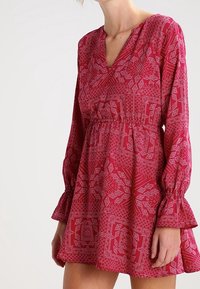 Rotes Kleid mit langen, weit ausgestellten Ärmeln, einem V-Ausschnitt und einem elastischen Bund. Es hat aufwendige weiße geometrische Muster überall.