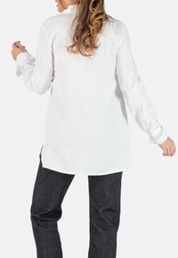 Witte blouse met lange mouwen en hoge kraag, gladde textuur, zijsplitten en knoopmanchetten, gecombineerd met donkere spijkerbroeken.