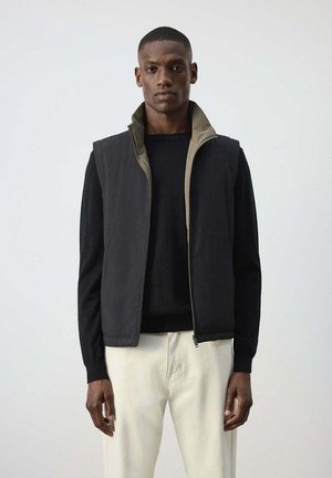 Hombre vestido con camisa negra de manga larga, chaleco negro con cremallera y forro beige, y pantalones beige claros, de pie frente a un fondo gris liso.