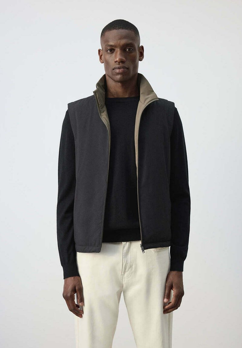 Homme portant une chemise noire à manches longues, un gilet noir zippé avec une doublure beige et un pantalon beige clair, se tenant devant un fond gris uni.