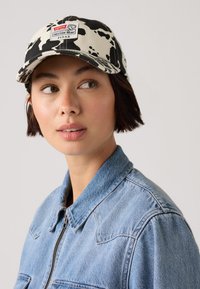 Czapka w print w krowie łatki z płaskim daszkiem, posiada naszywkę z logo. Noszona z jasnoniebieską kurtką dżinsową, która ma gładką teksturę i widoczne szwy.