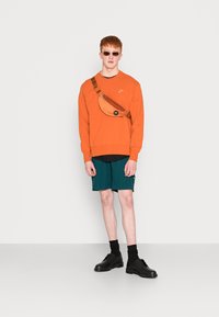 Oranger Sweatshirt mit schwarzem Logo, grüne Shorts und schwarze Schuhe. Zubehör umfasst eine braune Umhängetasche und Sonnenbrille.
