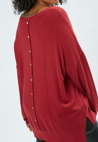 Roter, übergroßer Pullover aus weichem Strickmaterial. Verfügt über einen tiefen Rückenaufschneider mit dekorativen Knöpfen entlang der Mitte, langen Ärmeln und seitlichen Schlitzen.