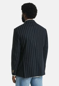 Il blazer a righe navy presenta un design sartoriale, spalle strutturate e dettagli con bottoni dorati. La fessura sul retro consente una maggiore libertà di movimento.