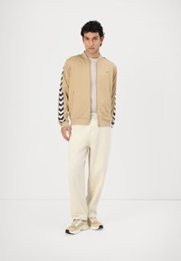 Beige bomber jacket with navy chevron stripes on sleeves, paired with cream pants and light-colored sneakers. Textured material, zipper front.
