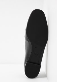 Chaussure en cuir noir avec une texture lisse et un bout arrondi. Le talon bas présente une surface texturée. Marquage visible sur la semelle.