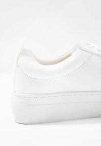 Vit lädersneaker med häl och dragtab samt tjock vit gummisul, visad mot en vit bakgrund.