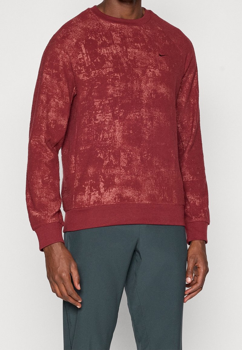 Homme portant un sweat-shirt rouge texturé à manches longues avec un petit logo Nike noir sur la poitrine et un pantalon vert foncé.