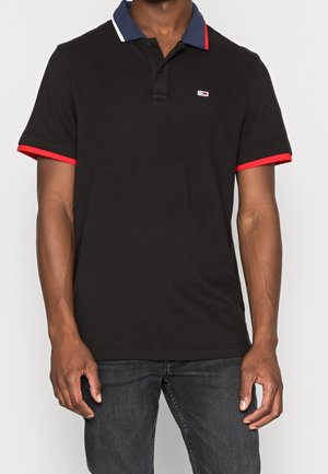 Homme portant un polo noir avec un col bleu marine et un bord de manche rouge, associé à un jean gris foncé, debout devant un fond blanc.