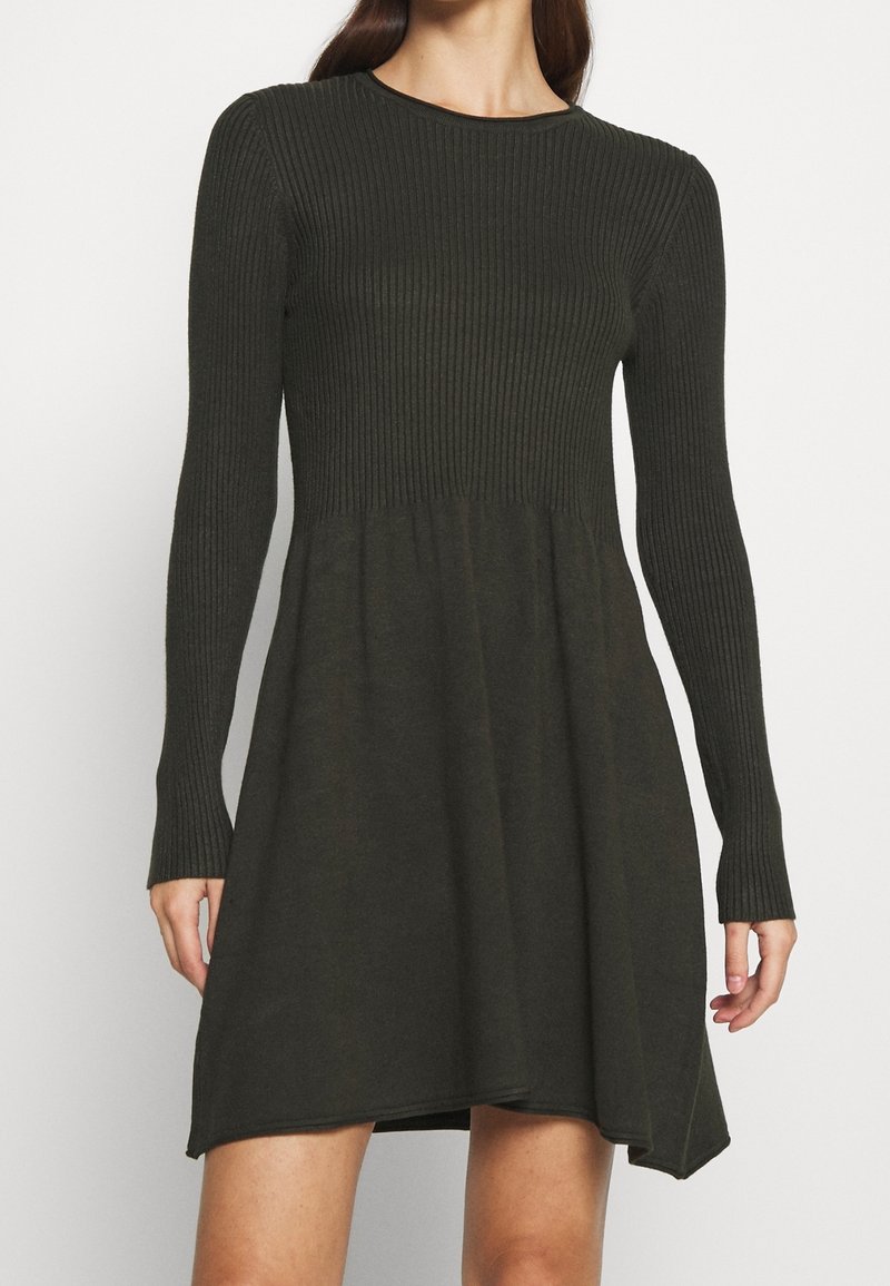 Femme portant une robe en maille côtelée vert foncé avec manches longues et une jupe légèrement évasée au-dessus des genoux.