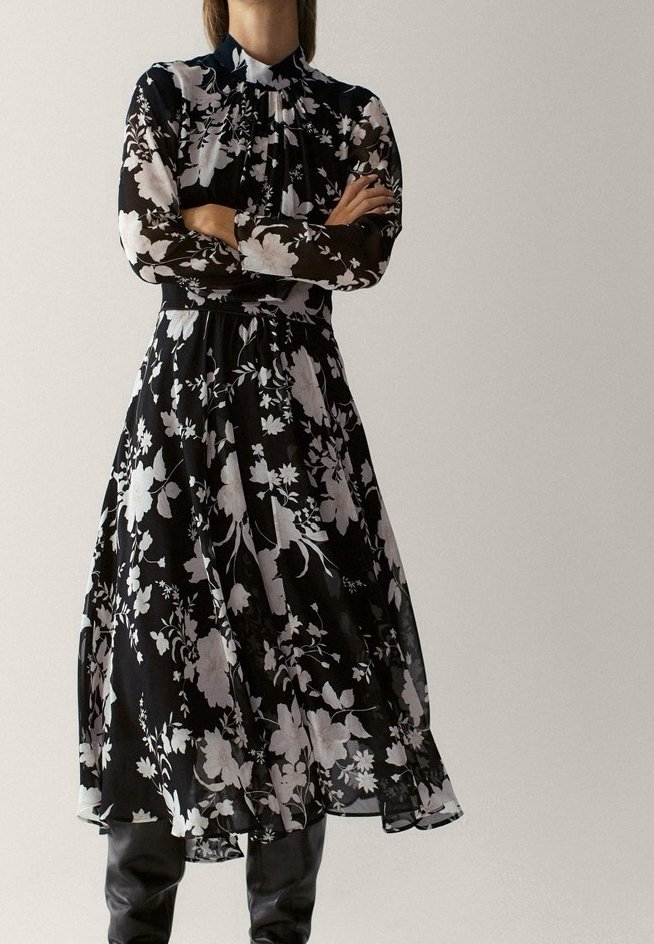 Femme portant une robe midi noire à motifs floraux blancs et manches longues, debout les bras croisés, assortie à des bottes noires montant jusqu'aux genoux.