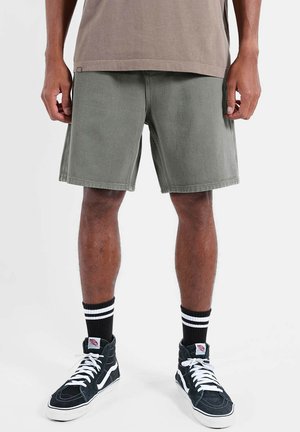 Homme portant un short vert olive, des baskets montantes noires avec des lacets blancs, et des chaussettes noires avec deux rayures blanches, debout sur un fond blanc.