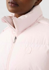 Lichtroze gewatteerde jas met hoge kraag, zilveren ritssluiting en gebosseleerd logo op de borst. Het materiaal lijkt glad en zacht van textuur.