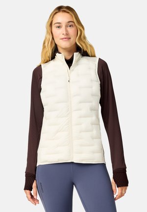Ung kvinde med langt blondt hår iført en hvid quiltet vest over en mørk langærmet skjorte og blå leggings mod en ensfarvet baggrund.