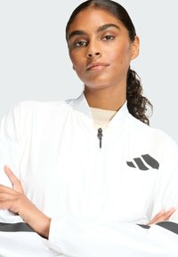 Bijela bomber jakna s visokim ovratnikom, crnim detaljima i logotipom. Glatka tekstura s patentnim zatvaračem. Model pozira prekriženih ruku.