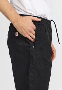 Hand in de zak van zwarte broek met elastische tailleband en ritssluiting, persoon draagt een wit shirt tegen een effen achtergrond.