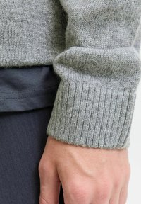 Poignet de pull en tricot gris avec une texture côtelée, associé à un tissu foncé en dessous, mettant en valeur un superposition sans couture et une douceur.