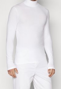 Mann in einem enganliegenden weißen Rollkragenpullover mit langen Ärmeln, kombiniert mit weißen Hosen, vor einem schlichten Hintergrund stehend.