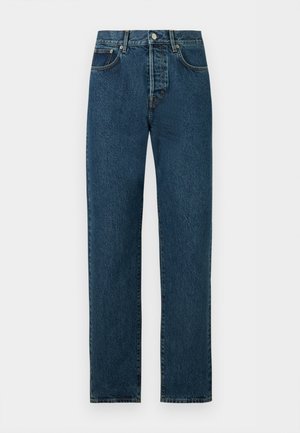 Ung mand med krøllet hår iført en åben hvid kortærmet skjorte over en hvid t-shirt, blå jeans, sort bælte og sorte lædersko, stående.