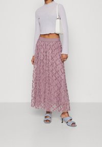 Ljust lila långärmad topp i kombination med en rosa, lager-på-lager, genomskinlig kjol med blommönster. Modellen har på sig blå öppna klackskor.