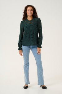 Dark green lace blouse with long sleeves, keyhole neckline, intricate floral patterns, paired with light blue jeans and black heeled shoes.