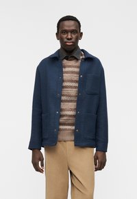 Μπουφάν από μαλλί σε ναυτικό μπλε χρώμα με δύο μπροστινές τσέπες και κουμπιά τύπου κλιπ, φορεμένο πάνω από ριγέ πλεκτό πουλόβερ και συνδυασμένο με μπεζ παντελόνι από βελουτέ υλικό.