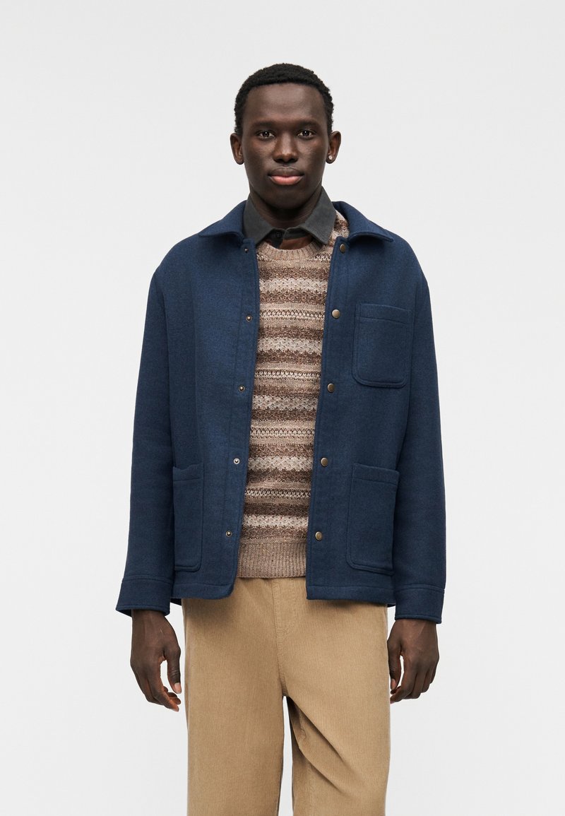 Μπουφάν από μαλλί σε ναυτικό μπλε χρώμα με δύο μπροστινές τσέπες και κουμπιά τύπου κλιπ, φορεμένο πάνω από ριγέ πλεκτό πουλόβερ και συνδυασμένο με μπεζ παντελόνι από βελουτέ υλικό.