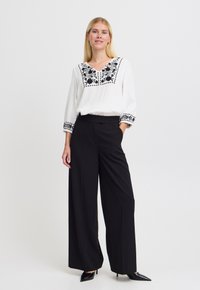 Blusa bianca ricamata con motivi floreali neri, abbinata a pantaloni neri a gamba larga e tacchi a punta neri. Tessuto morbido, vestibilità rilassata.