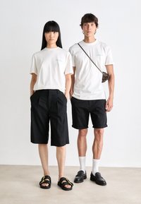 Two models wear neutral-colored t-shirts, black shorts, and sandals. The woman has loose shorts; the man has tailored shorts and loafers.