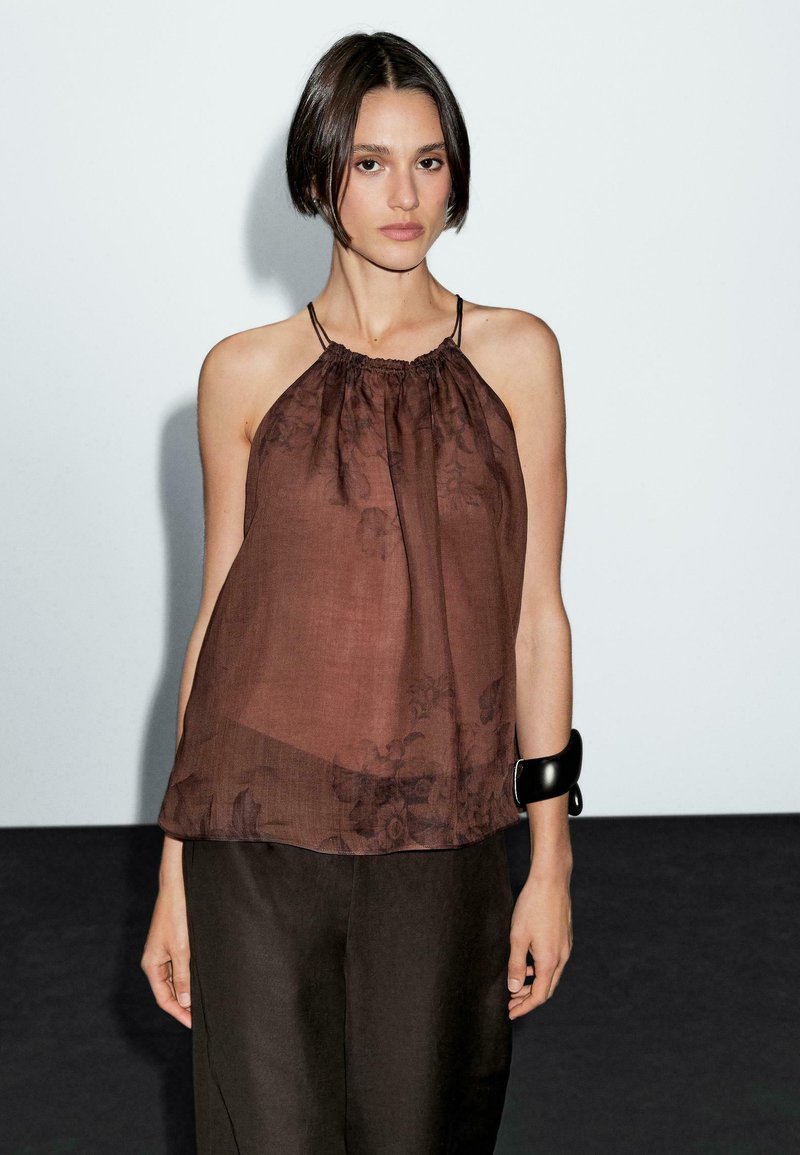Top halter trasparente marrone con scollo arricciato e motivo floreale. Indossato con pantaloni larghi marrone scuro e un bracciale nero chunky.