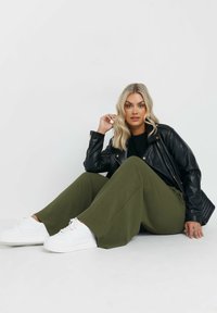 Svart läderjacka, svart topp, olivgröna vida byxor och vita sneakers. Modellen sitter med handen på knäet, strukturerad tyg synlig.