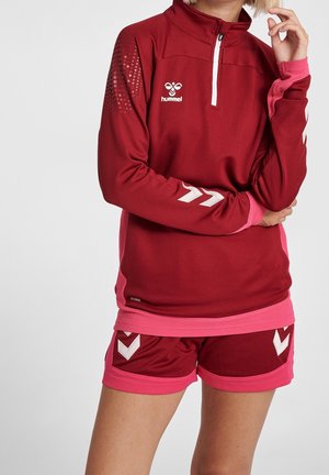 Osoba ubrana w bordowo-różowy strój sportowy hummel, w tym bluzę z długim rękawem z półzamkiem i pasujące spodenki, pozująca ze skrzyżowanymi ramionami.