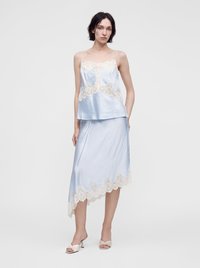 Femme portant un caraco en satin bleu clair et une jupe asymétrique avec un bord en dentelle crème, associée à des sandales blanches à talons ouverts, debout.