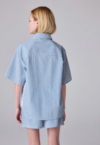 Pamucna prugasta košulja u svijetlo plavoj i bijeloj boji, kratkih rukava, ležernog kroja, s yoke-om na leđima i bočnim prorezom za prozračivanje.