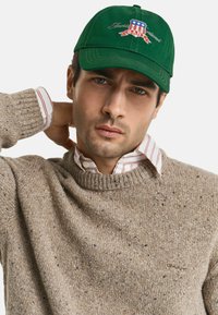 Mann in beigem, gesprenkeltem Strickpullover und gestreiftem Hemd mit Kragen, trägt grüne Mütze mit rot-weiß-blauem Emblem, Hand hinter dem Nacken.