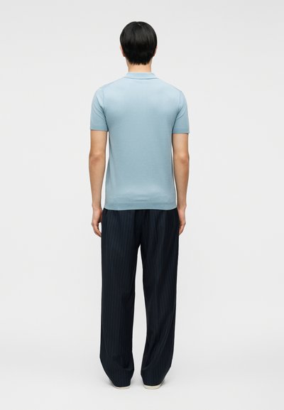 Άνδρας που στέκεται πλάτη φοράει ανοιχτό μπλε πολο με κοντά μανίκια και φαρδιά μαύρη ριγέ παντελόνια μπροστά σε απλό φόντο.