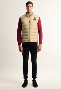 Chaleco acolchado beige con cremallera frontal, llevado sobre un suéter burdeos, combinado con jeans negros y zapatos marrones. Presenta parches de logo en el chaleco.