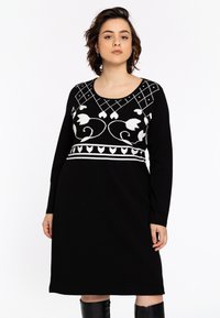Modèle portant une robe noire à manches longues avec des broderies florales blanches et des motifs géométriques, se tenant avec confiance et une expression neutre.