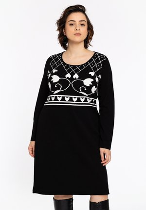 Modèle portant une robe noire à manches longues avec des broderies florales blanches et des motifs géométriques, se tenant avec confiance et une expression neutre.