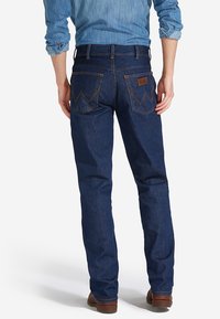 Jeans de mezclilla azul oscuro con pierna recta. Presenta dos bolsillos traseros con costuras en zigzag y una etiqueta de cuero marrón en la cintura.