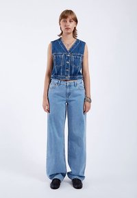 Spijkervest met knoopsluiting aan de voorkant, lichtblauwe oversized jeans en zwarte schoenen. Kenmerken zijn een hoge taille en een wijd uitlopende pijp.