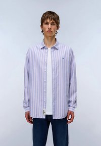 Chemise à rayures boutonnée violet clair et blanc, avec manches longues, coupe décontractée, et petite étiquette logo sur l'ourlet. Associée à un jean bleu.