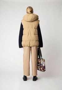 Befodrad beige väst med en stor huva, kombinerad med lös sittande beige byxor och en mönstrad väska. Svarta kraftiga stövlar fulländar outfiten.