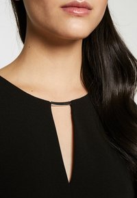 Svart klänning med V-ringning, med en silverdetalj vid kragen. Slät textur, närbild av hals- och axelområdet.