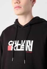 Svart hettegenser med hvit grafikk "CALVIN KLEIN" logo og røde detaljer. Har snorer med metallspisser og en teksturert stoff.